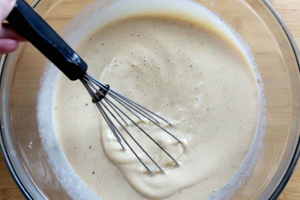 A hand holding a whisk is mixing a creamy, light brown batter in a clear glass bowl on a wooden surface.