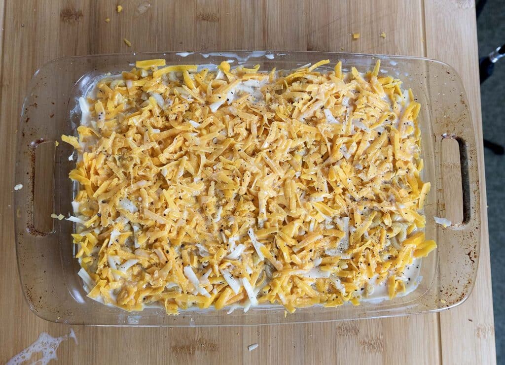A glass baking dish filled with a mixture topped with shredded yellow and white cheese and ground pepper, sitting on a wooden surface.