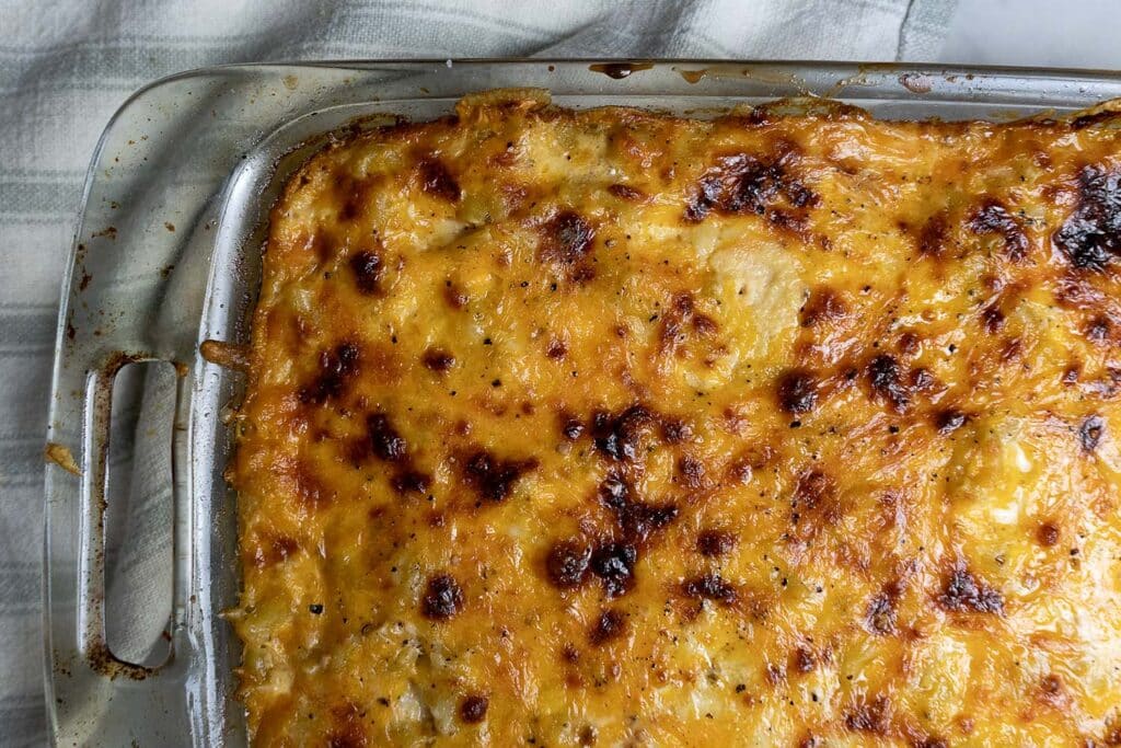 A close-up view of a baked casserole with a golden, bubbly, and slightly browned cheese topping in a glass dish, resting on a striped cloth.