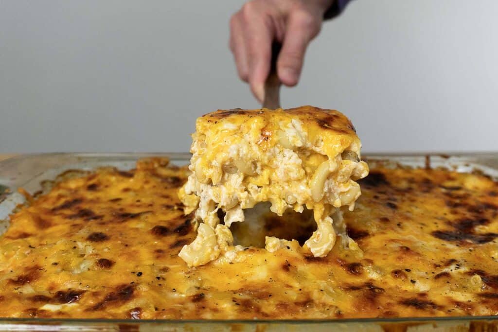 A hand holds a spatula lifting a square serving of baked macaroni and cheese from a glass dish, showing gooey melted cheese and golden-brown crust.