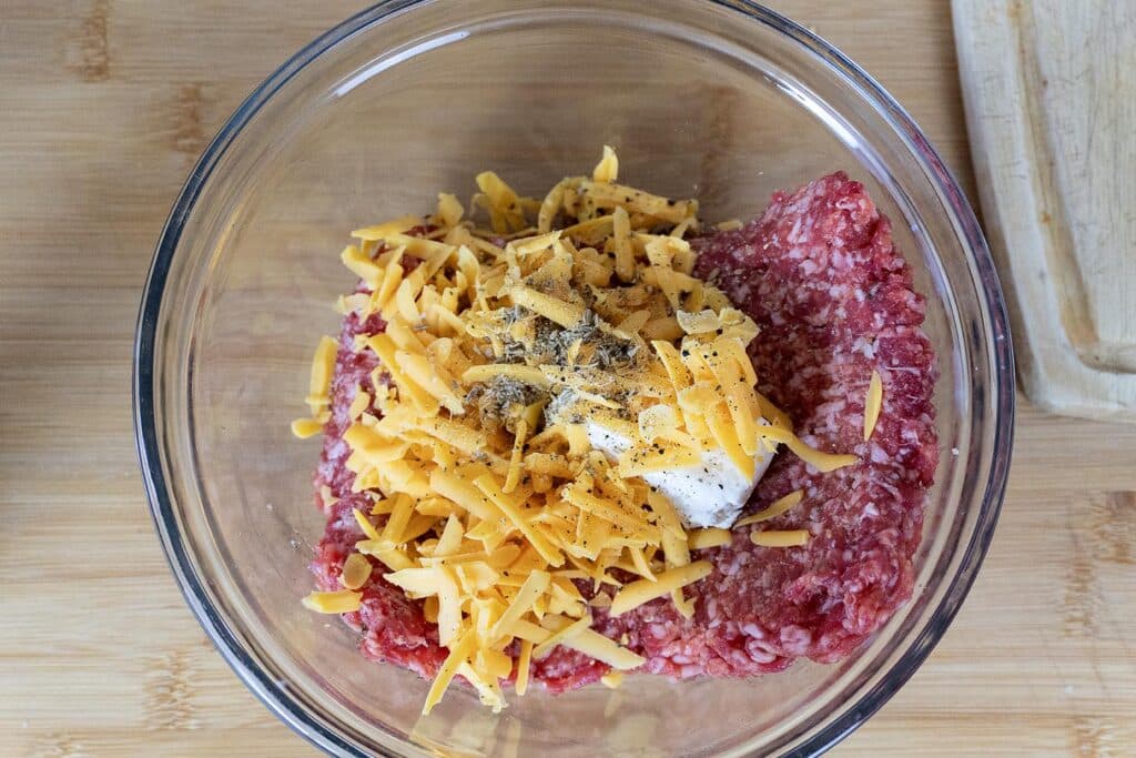 A glass bowl containing raw ground meat, shredded cheddar cheese, a dollop of white sauce, and black pepper, placed on a wooden surface next to a cutting board.
