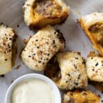 A plate with several pieces of puff pastry sausage rolls topped with sesame and poppy seeds, served with a small dish of creamy dipping sauce.