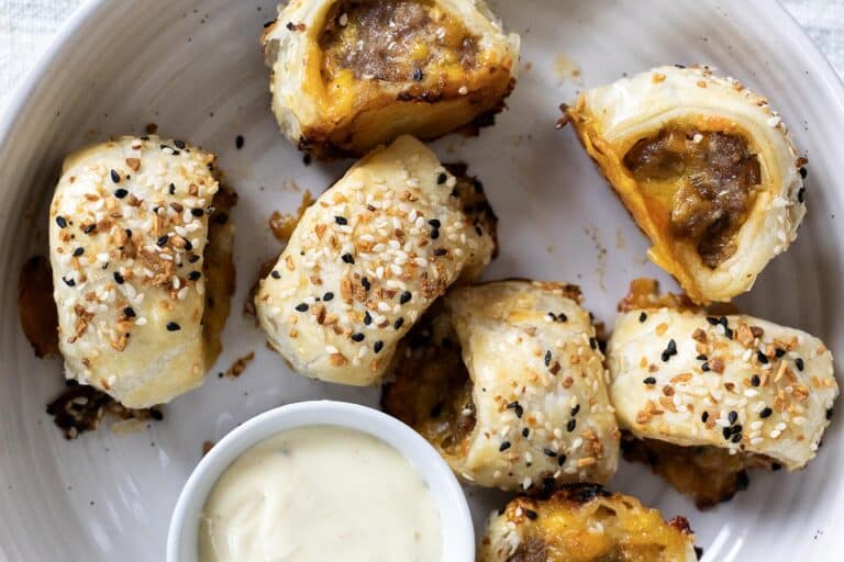 A plate with several pieces of puff pastry sausage rolls topped with sesame and poppy seeds, served with a small dish of creamy dipping sauce.