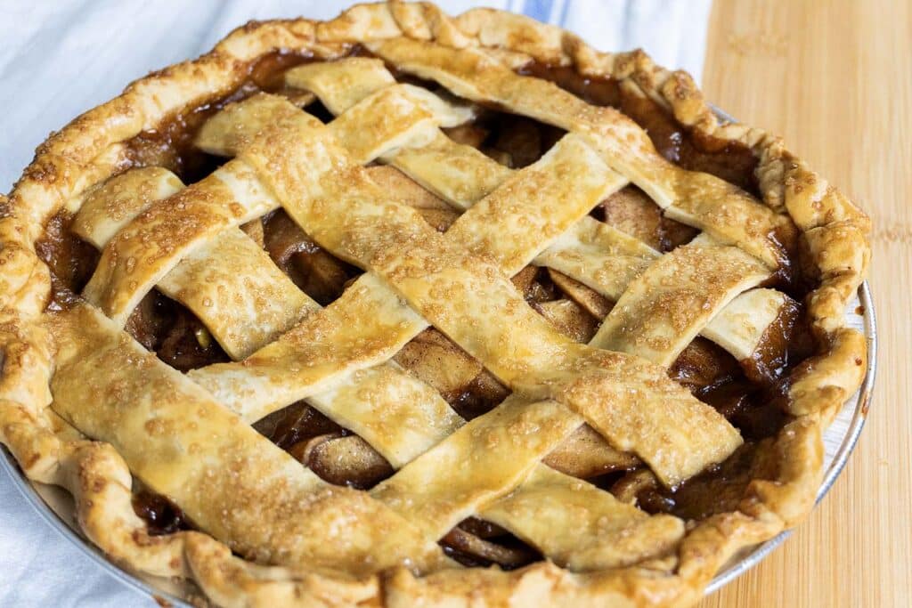A golden-brown apple pie with a lattice crust sits on a wooden surface, its filling visible between the woven pastry strips. Sugar crystals are sprinkled on top, and the edge of the pie is crimped.