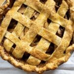 A freshly baked apple pie with a golden-brown lattice crust sits on a white towel with a blue stripe, showing bubbly filling and sugar sprinkled on top.