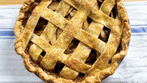 A freshly baked apple pie with a golden-brown lattice crust sits on a white towel with a blue stripe, showing bubbly filling and sugar sprinkled on top.