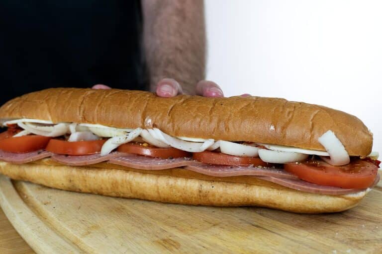 A person holds a long sub sandwich on a wooden board, filled with layers of sliced ham, tomato, and onions inside a fresh hoagie roll.