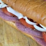 Close-up of a sub sandwich on a wooden surface, filled with slices of salami, tomato, onion, and cheese inside a soft hoagie roll.