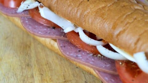 Close-up of a sub sandwich on a wooden surface, filled with slices of salami, tomato, onion, and cheese inside a soft hoagie roll.
