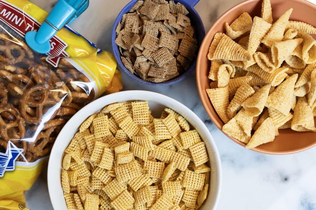 Four bowls and bags contain different snack foods: pretzels, brown and yellow Chex cereals, and corn chips, arranged on a marble surface. A teal clip is attached to the pretzel bag.