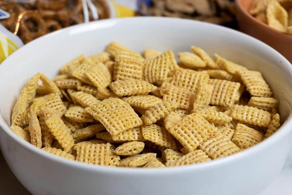 A white bowl filled with square-shaped, woven breakfast cereal pieces, with other snacks partially visible in the background.