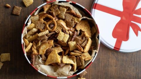 A festive tin with a red bow design holds a mix of Chex cereal, pretzels, nuts, and crackers on parchment paper. Some snack mix pieces are scattered on a wooden surface next to the tin.