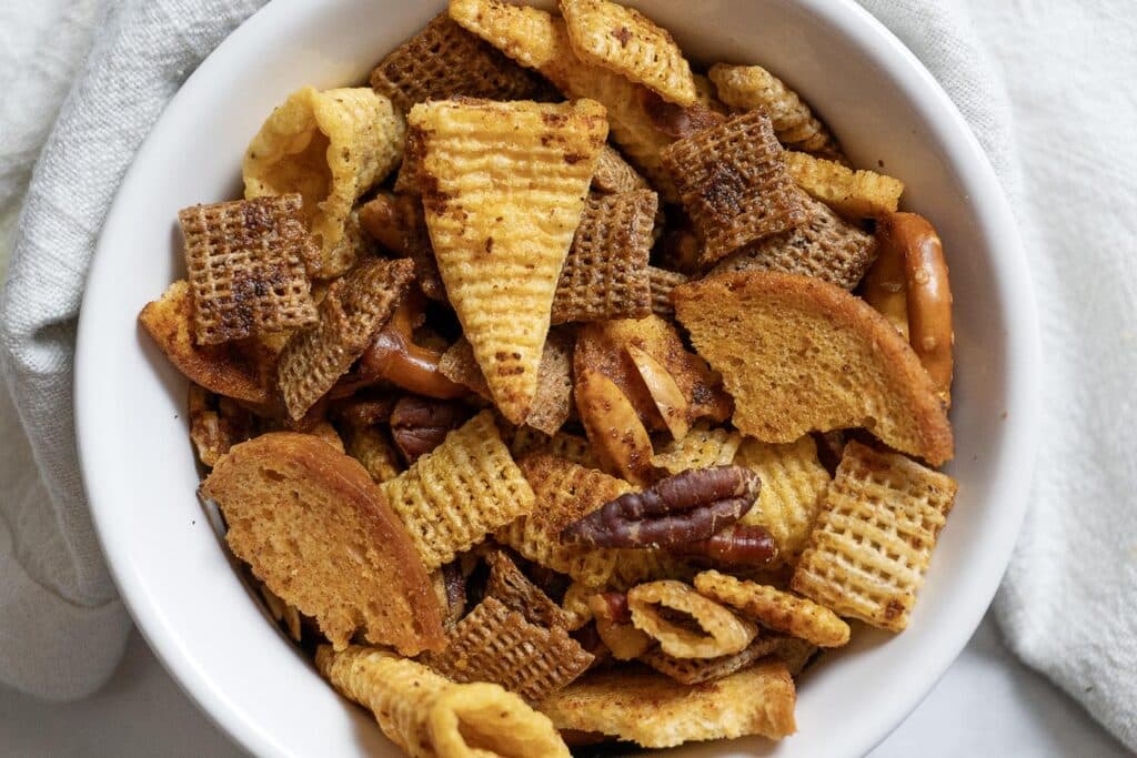 A white bowl filled with a snack mix including various types of cereal squares, bagel chips, pretzel sticks, corn chips, and pecans, placed on a light-colored cloth.