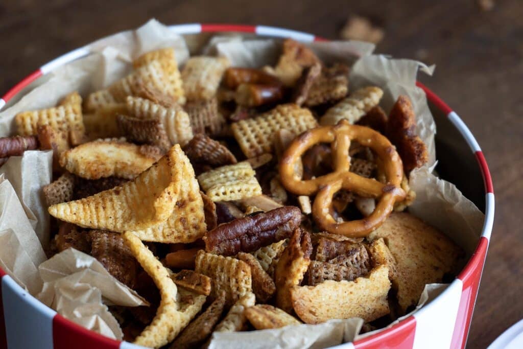 A close-up of a red and white tin filled with assorted snack mix, including pretzels, cereal squares, pecans, rye chips, and corn chips, all on crumpled parchment paper.