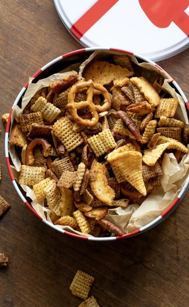 A round tin lined with parchment paper and filled with Chex mix snack, including pretzels, cereal squares, breadsticks, and crackers, sits on a wooden surface. The lid of the tin is partially visible.