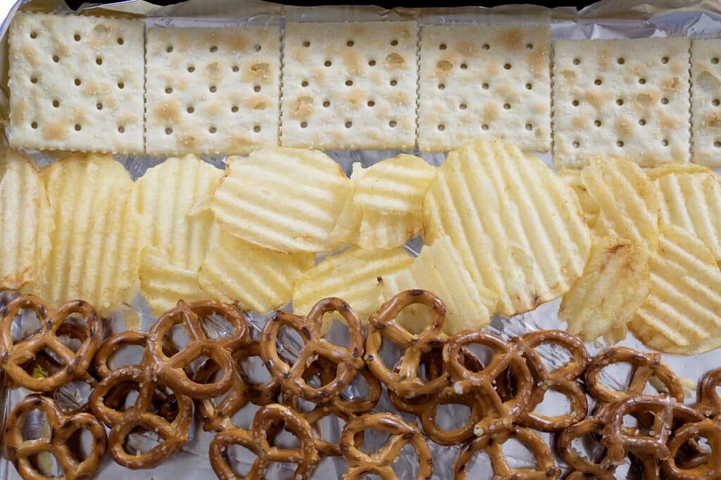 A neatly arranged row of saltine crackers, ridged potato chips, and pretzels sits on a sheet of foil, creating three distinct horizontal bands of snacks.