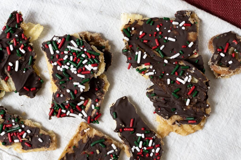Broken pieces of chocolate-covered potato chips topped with red, white, and green holiday sprinkles are scattered on a white cloth.