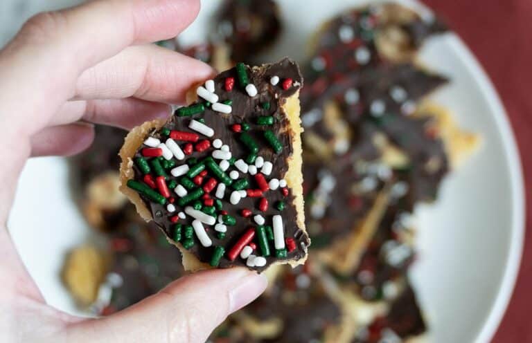 A hand holds a piece of chocolate-covered cookie topped with red, white, and green sprinkles, with more decorated cookies blurred in the background on a white plate.