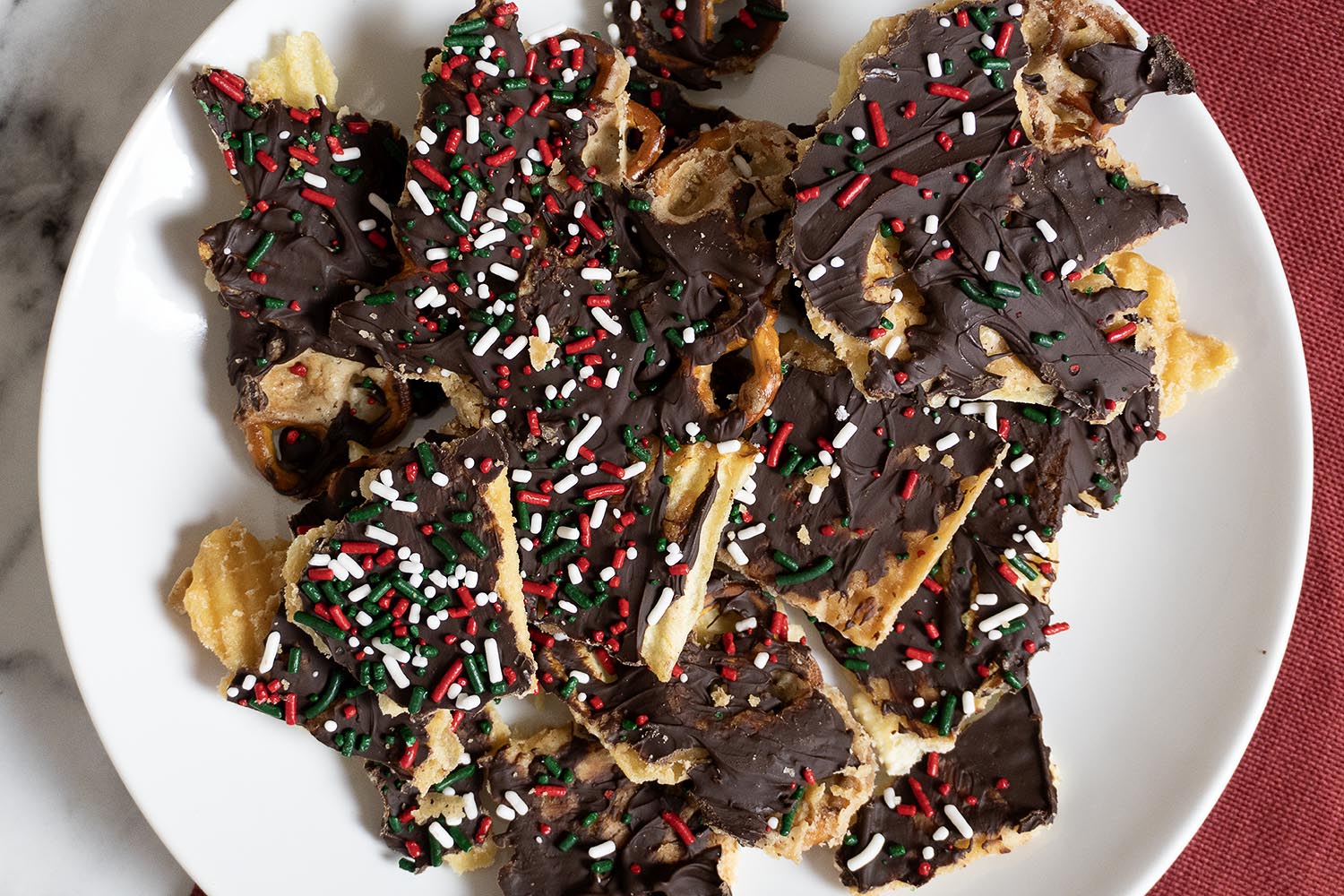 A white plate filled with pieces of chocolate-covered cracker bark topped with red, white, and green sprinkles, set on a red cloth background.