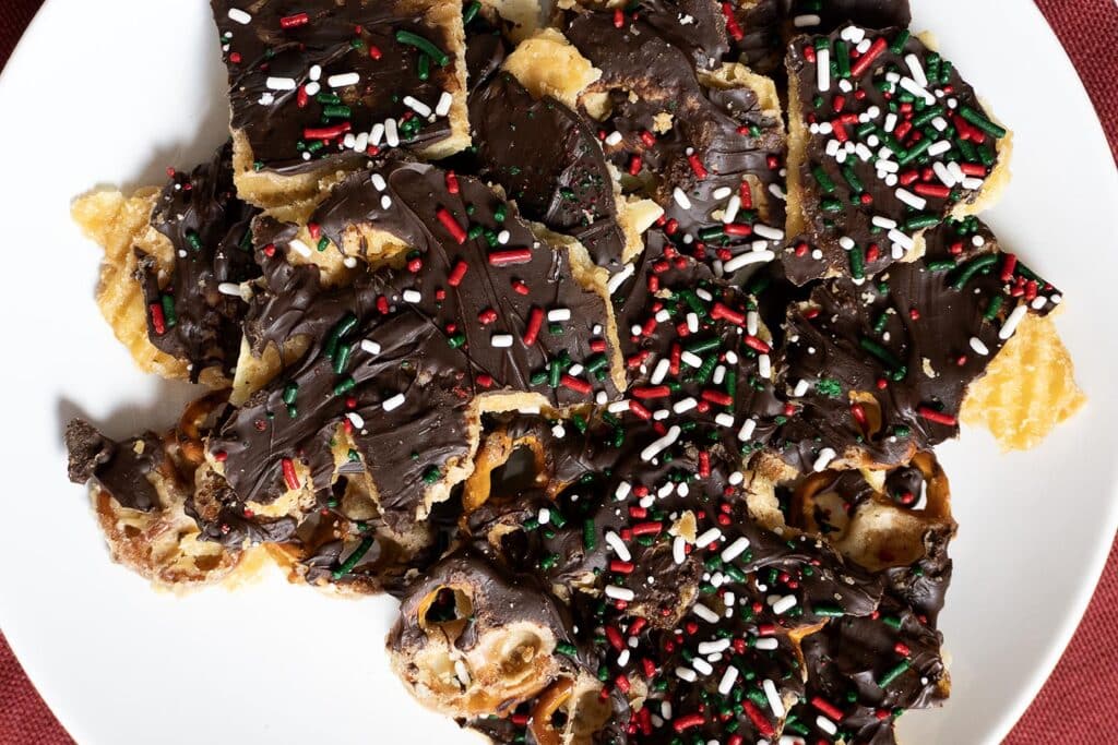A plate of chocolate-covered pretzel bark topped with red, white, and green sprinkles is arranged on a white dish.