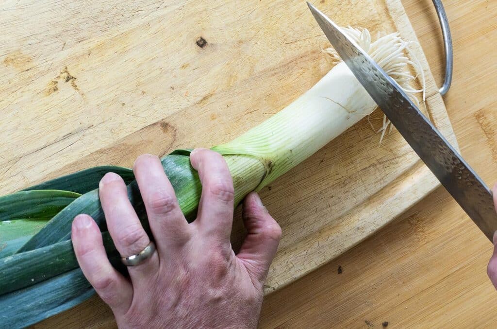 A person’s hand holds a leek on a wooden cutting board while slicing off the root end with a large knife.