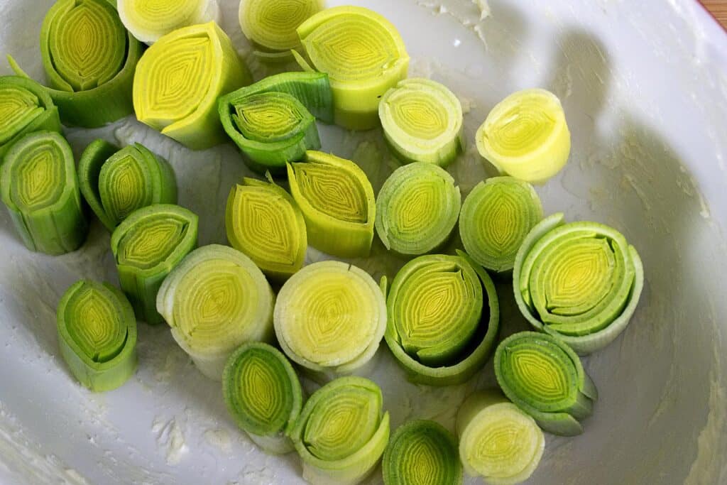 Sliced leeks, showing light green and yellow centers, are arranged in a white dish. The concentric rings and varying shades give a fresh, vibrant appearance.