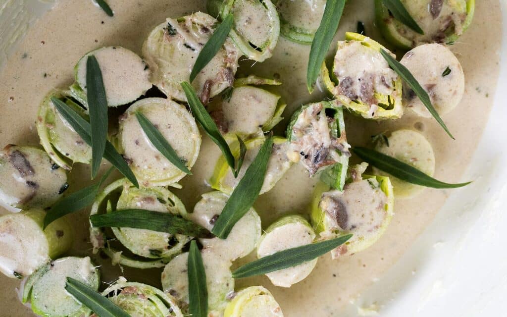 Sliced leeks in a creamy brown sauce, garnished with fresh green herbs, are arranged on a white plate. The sauce contains visible bits of mushroom or similar ingredients.