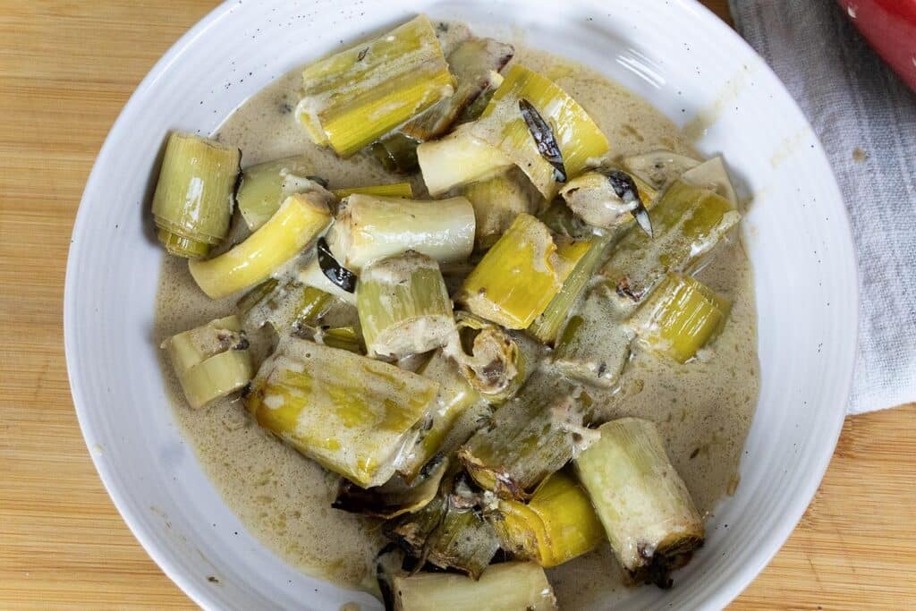A white bowl filled with chopped cooked leeks in a creamy sauce, set on a wooden surface with a white napkin partially visible to the side.
