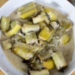 A white bowl filled with chopped cooked leeks in a creamy sauce, set on a wooden surface with a white napkin partially visible to the side.