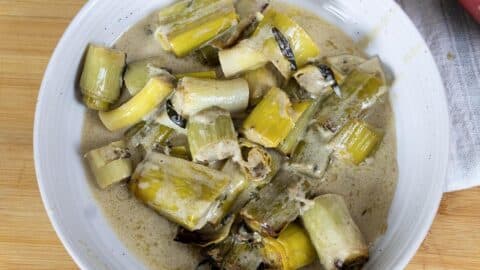 A white bowl filled with chopped cooked leeks in a creamy sauce, set on a wooden surface with a white napkin partially visible to the side.