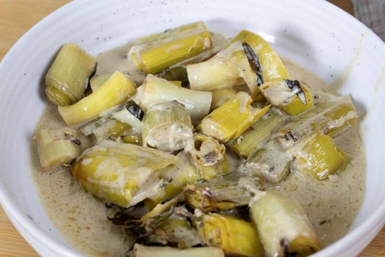 A white bowl filled with creamy braised leeks, cut into chunks and coated in a light sauce, with some browned and caramelized edges visible.