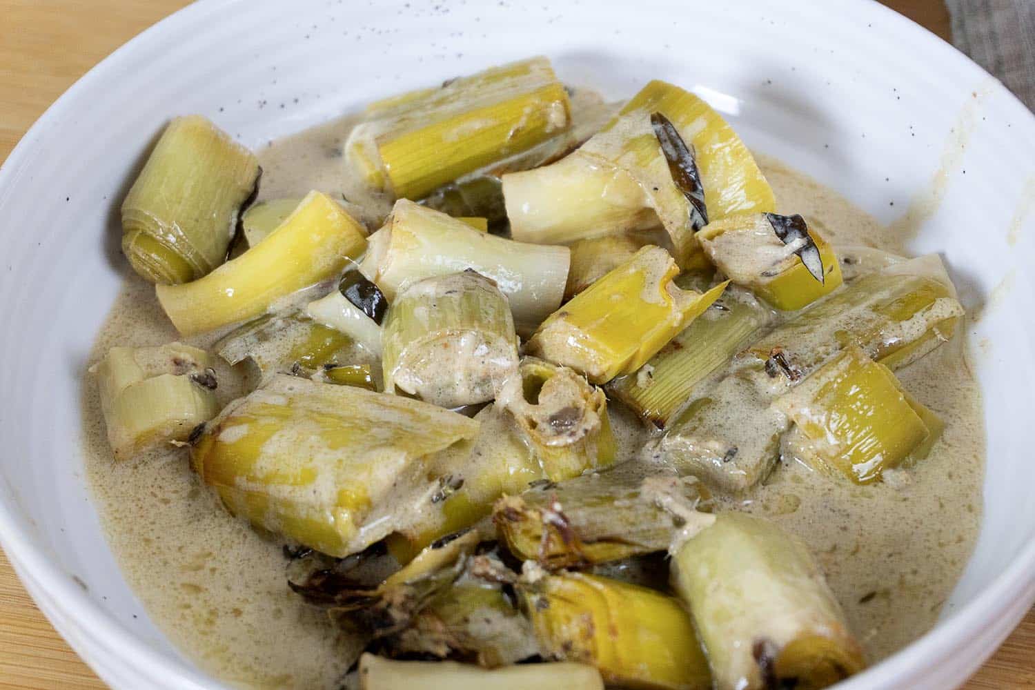 A white bowl filled with creamy braised leeks, cut into chunks and coated in a light sauce, with some browned and caramelized edges visible.