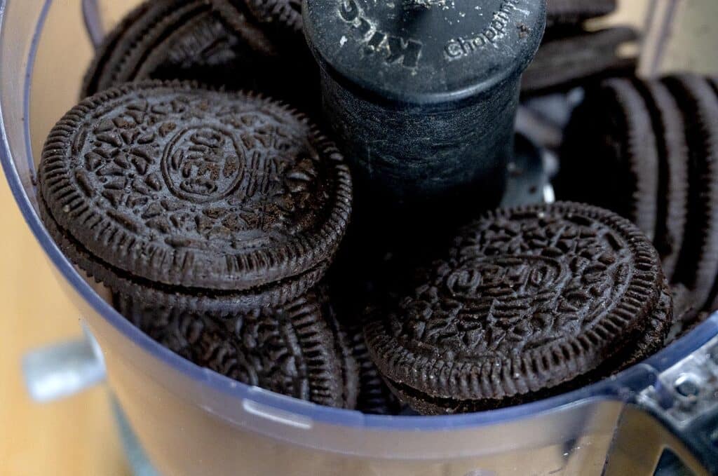A close-up view of several chocolate sandwich cookies with cream filling inside a food processor, ready to be crushed.