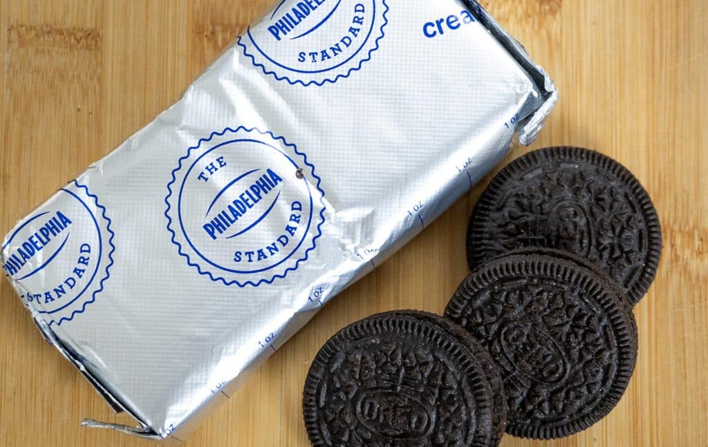 A block of Philadelphia cream cheese in silver packaging sits on a wooden surface next to three chocolate sandwich cookies with cream filling.