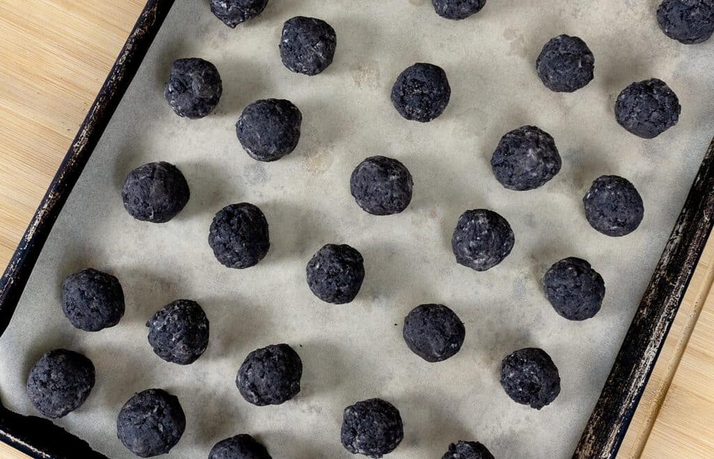 A baking tray lined with parchment paper holds evenly spaced, round, dark gray or black dough balls, ready to be baked. The tray rests on a light wooden surface.