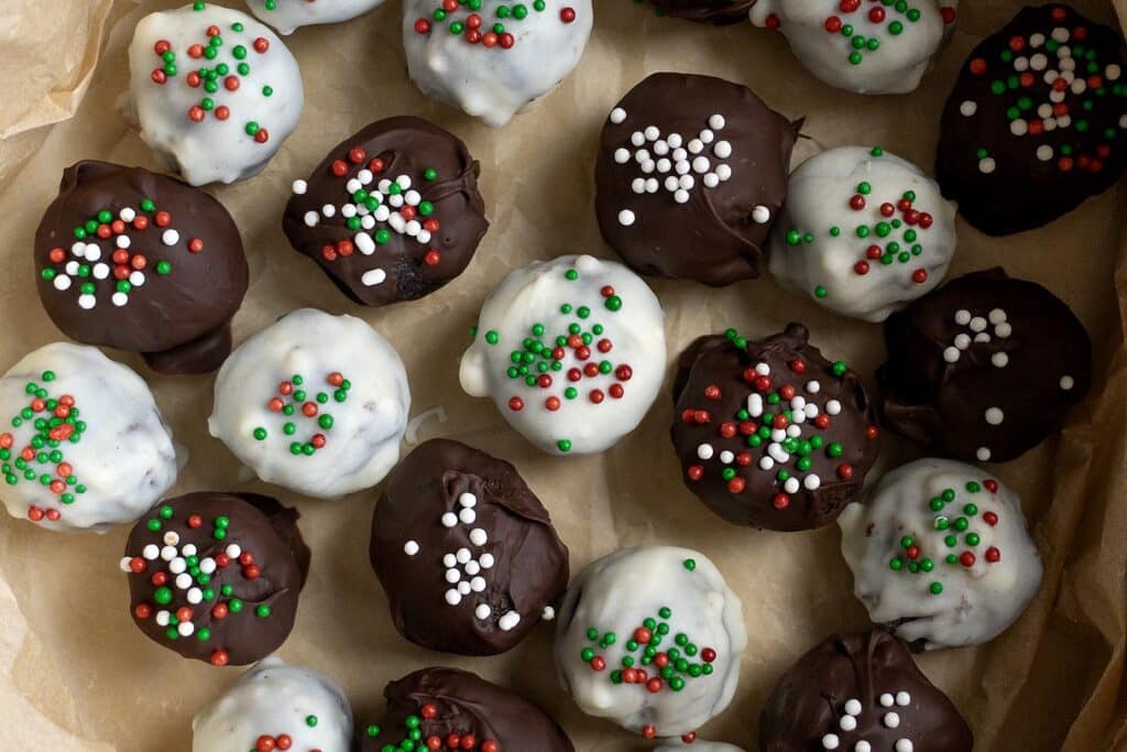 A batch of round chocolate truffles coated in white and dark chocolate, topped with red, green, and white sprinkles, arranged on parchment paper.
