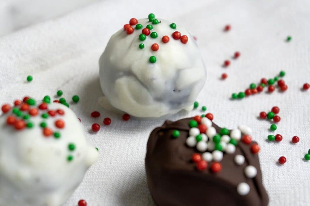 Three holiday-themed truffles on a white cloth, two coated in white chocolate and one in dark chocolate, all topped with red, green, and white sprinkles.