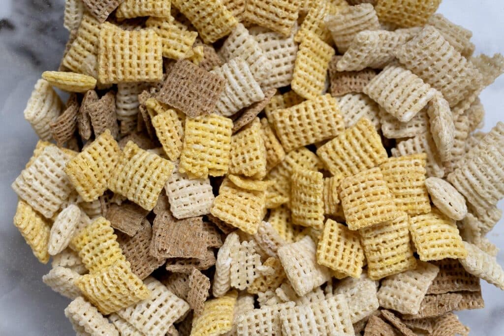 A close-up of mixed Chex cereal pieces in various colors—yellow, brown, and white—displaying their distinct square shapes and woven textures.