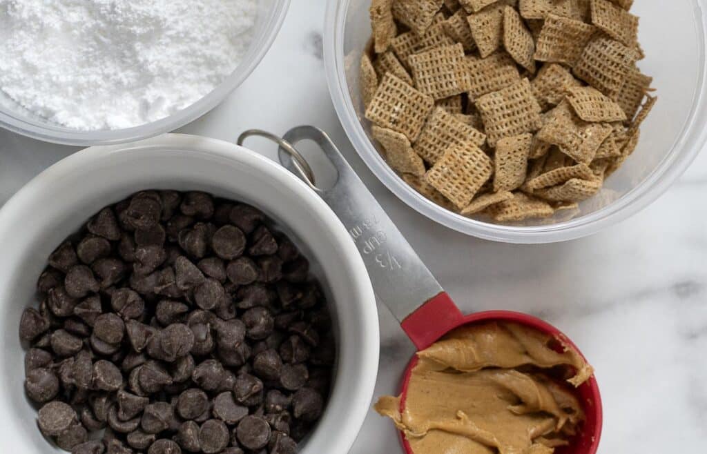 A top-down view of baking ingredients: powdered sugar, cereal squares, chocolate chips, and a measuring cup filled with peanut butter on a white surface.