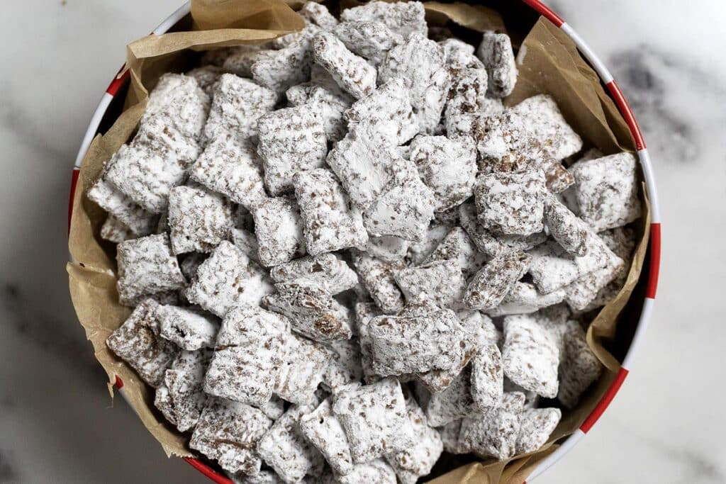 A bowl lined with brown parchment paper is filled with Chex cereal squares coated in powdered sugar, also known as puppy chow or muddy buddies, on a white marble surface.
