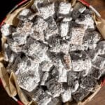 A round tin lined with parchment paper is filled with chocolate-covered cereal squares coated in powdered sugar, commonly known as puppy chow or muddy buddies, sitting on a wooden surface.