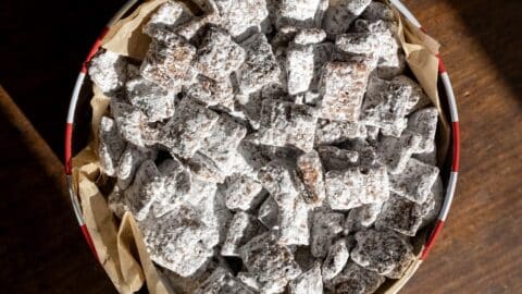A round tin lined with parchment paper is filled with chocolate-covered cereal squares coated in powdered sugar, commonly known as puppy chow or muddy buddies, sitting on a wooden surface.