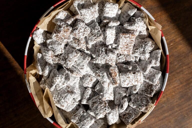A round tin lined with parchment paper is filled with chocolate-covered cereal squares coated in powdered sugar, commonly known as puppy chow or muddy buddies, sitting on a wooden surface.
