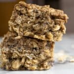 Two thick, square oat bars stacked on top of each other on a white surface, with visible oats and a chewy texture. A few scattered oats are in the background.