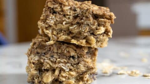 Two thick, square oat bars stacked on top of each other on a white surface, with visible oats and a chewy texture. A few scattered oats are in the background.