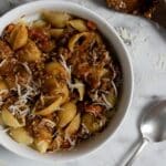 A white bowl filled with pasta shells topped with chunky meat and tomato sauce, garnished with shredded cheese. A spoon and a wooden spatula with sauce are on a marble surface beside the bowl.
