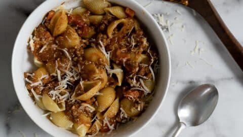 A white bowl filled with pasta shells topped with chunky meat and tomato sauce, garnished with shredded cheese. A spoon and a wooden spatula with sauce are on a marble surface beside the bowl.