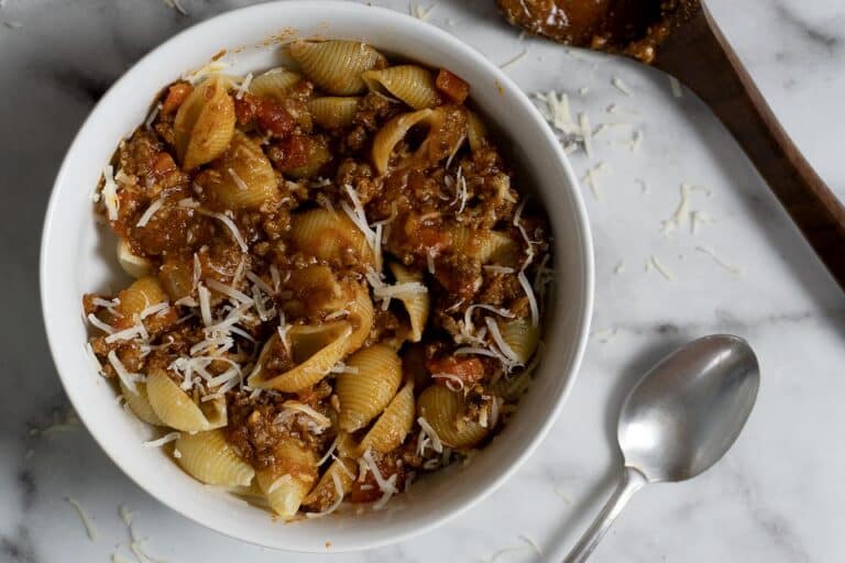 A white bowl filled with pasta shells topped with chunky meat and tomato sauce, garnished with shredded cheese. A spoon and a wooden spatula with sauce are on a marble surface beside the bowl.