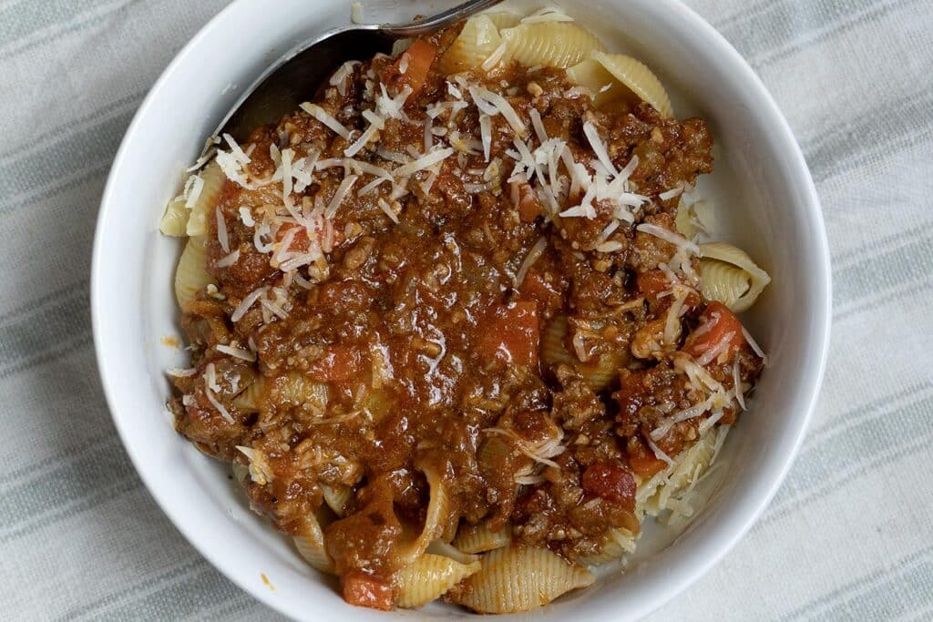 A white bowl filled with pasta shells topped with a chunky meat and tomato sauce, garnished with shredded cheese, sits on a light striped cloth.