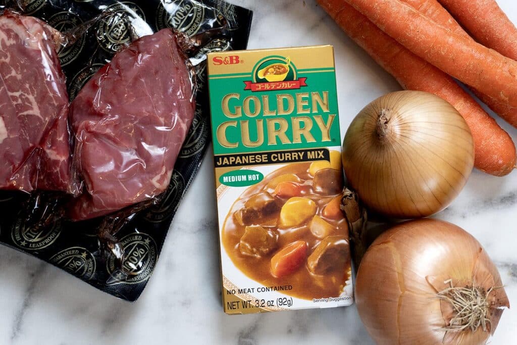 Vacuum-sealed beef, a box of S&B Golden Curry Japanese curry mix, two yellow onions, and several carrots are arranged on a white marble surface.